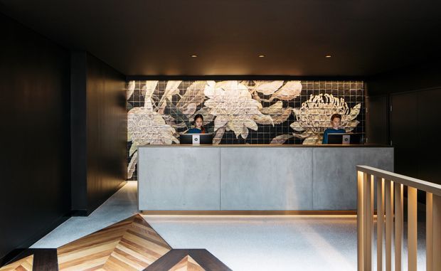 A dark, sophisticated reception lobby with two reception staff behind a large pale stone or concrete-fronted desk. The space features black walls and ceiling, warm recessed lighting, a dramatic botanical mural behind the desk, and a timber chevron-patterned floor or seating element in the foreground. A modern metal balustrade frames the right side.