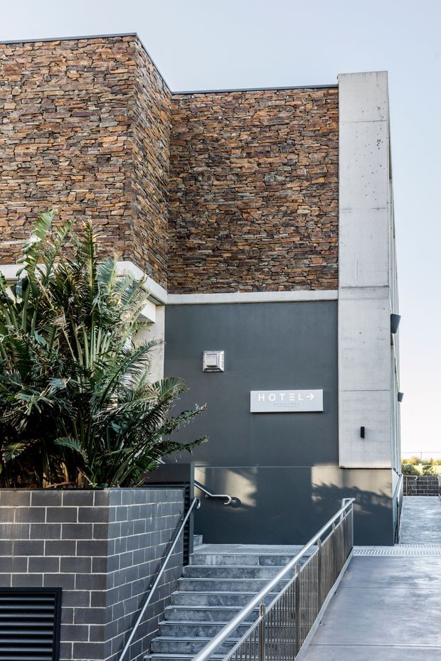 Exterior architectural photograph of a contemporary commercial building entrance. The facade combines stacked natural stone cladding, dark painted wall surfaces, exposed concrete vertical elements, and dark masonry planter walls. A stair and accessible ramp with metal handrails lead to the entrance, which is marked by a hotel sign. Tropical planting occupies the foreground.