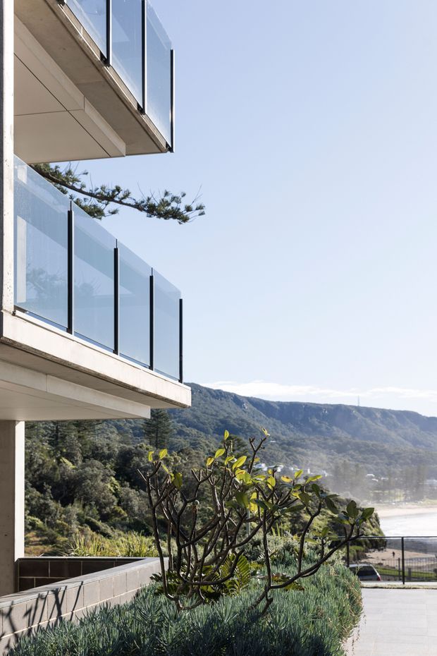 Exterior architectural view of a contemporary multi-level building with projecting concrete balconies and frameless glass balustrades. A landscaped terrace with drought-tolerant planting occupies the foreground, while a mountainous coastal or valley landscape is visible beyond.