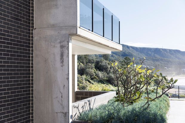 Exterior architectural view of a contemporary building with dark brick cladding, exposed concrete structure, a projecting upper-level balcony with frameless glass balustrade, and a landscaped terrace. A mountainous coastal landscape is visible beyond the building.