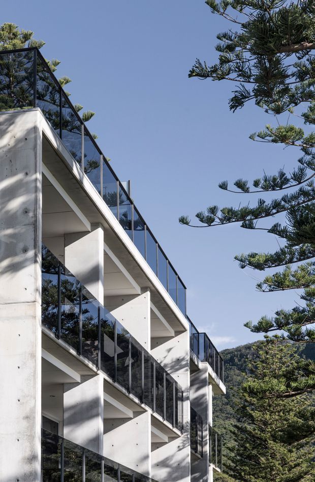 Exterior architectural photograph of a multi-storey modern concrete building with stacked projecting balconies, slim dark-framed glass balustrades, and clean geometric forms. Tall evergreen trees surround the structure, with a wooded hillside visible behind it under a clear blue sky.