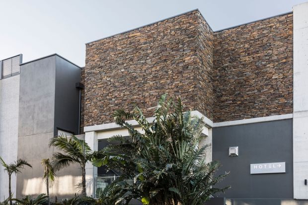 Exterior view of a contemporary hotel entrance or facade, featuring a dark rendered wall, large stacked natural-stone cladding panels, white structural trim, and tropical landscaping with palms in the foreground. A small hotel sign is visible on the wall.