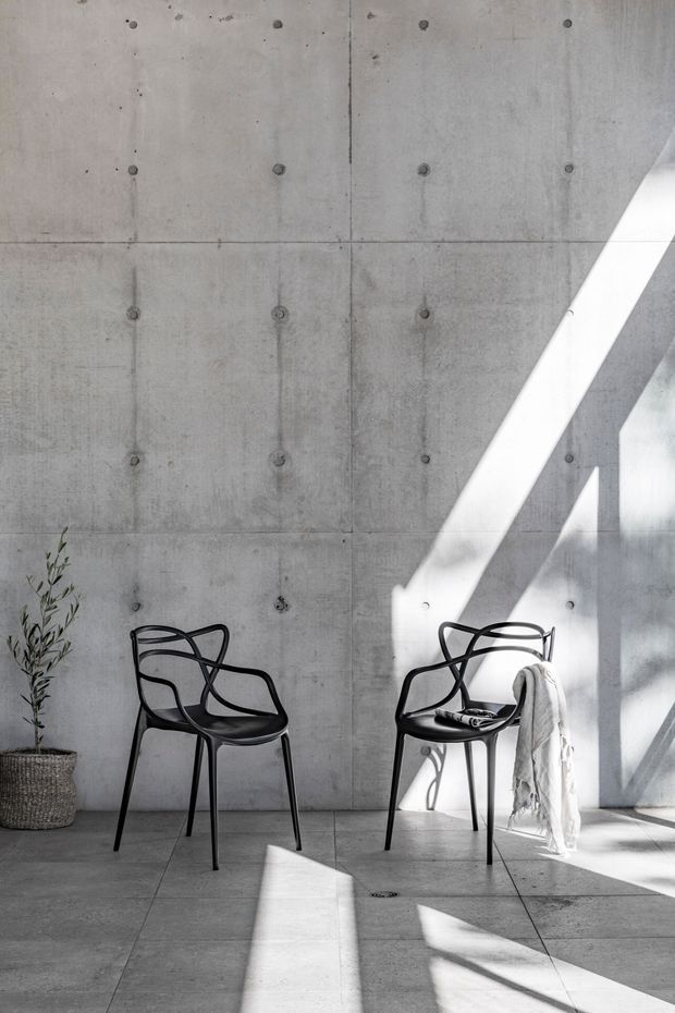 Minimalist interior with exposed, panel-formed concrete walls and matching concrete floor. Two black sculptural chairs sit in a sunlit space, accompanied by a small potted plant. Strong diagonal daylight creates pronounced shadows across the room.