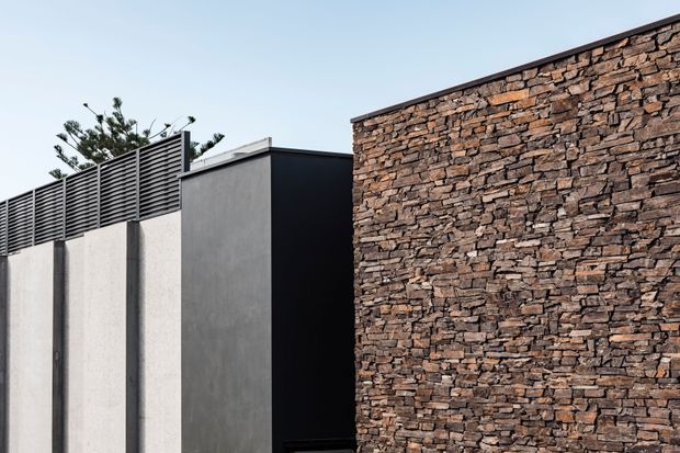 Exterior architectural detail showing a contemporary composition of smooth dark grey rendered wall panels, a black volume, horizontal louvred screening, and a prominent rugged stacked-stone feature wall. The image is tightly framed on the building facade against a clear sky, with minimal landscaping visible behind the screen.