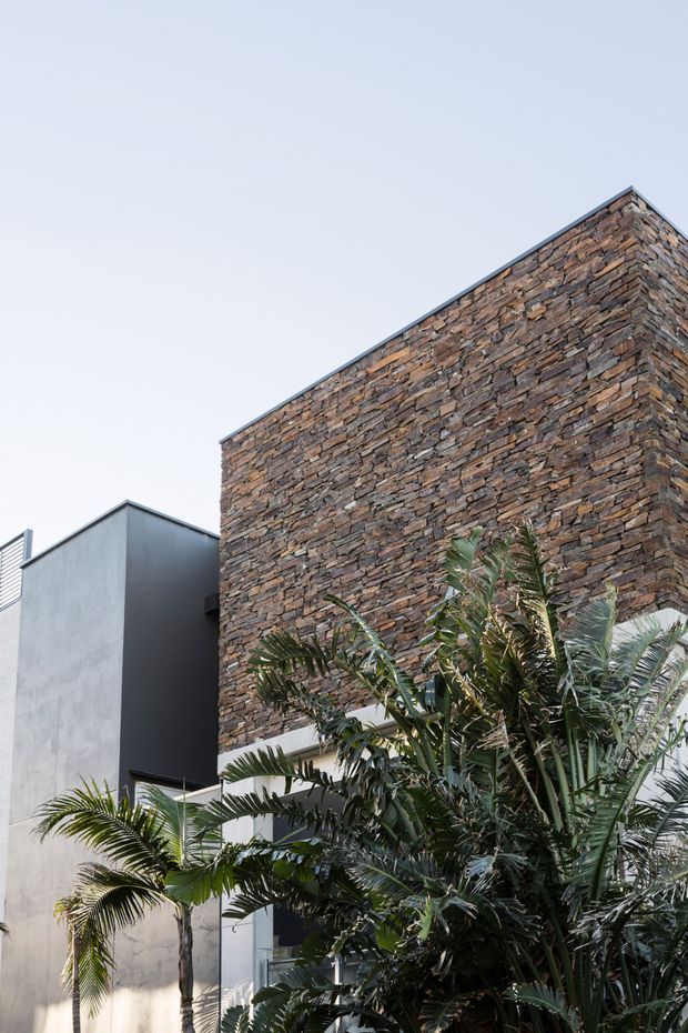 Exterior architectural view of a contemporary building with a tall facade clad in irregular stacked natural stone, paired with smooth rendered grey walls and a flat roofline. Tropical palms and dense planting partially obscure the lower facade.