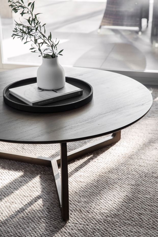 A close-up interior detail of a round dark wood coffee table with slender angled legs, positioned on a textured light grey carpet. A black circular tray holding a white ceramic vase with leafy branches and a design book sits on the tabletop. Strong sunlight creates shadows across the softly styled, neutral-toned living area.