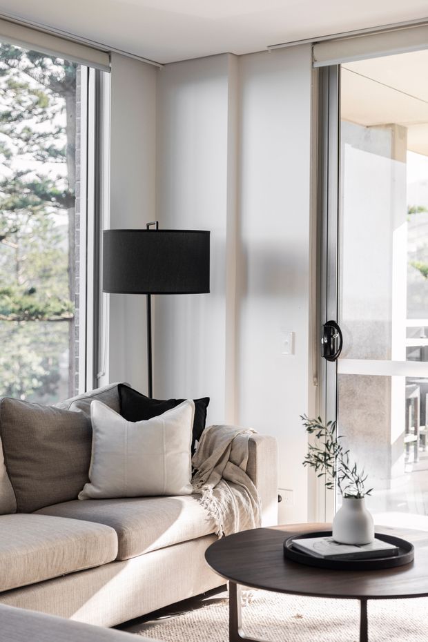Bright, softly styled living room with a large neutral upholstered sofa, layered cushions and throw, black floor lamp, round dark coffee table, and a small vase with greenery. Full-height windows and a glazed door bring in abundant natural light and overlook trees and a neighbouring structure. The palette is restrained in white, beige, grey, and black with a calm contemporary feel.