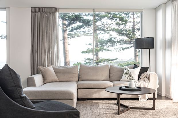 Bright contemporary living room with a large neutral L-shaped sectional sofa, dark lounge chair, round black coffee table, floor-to-ceiling windows, and full-height curtains. Coastal trees and water are visible beyond the glazing, while the room features light walls, a textured neutral rug, and restrained decorative styling.