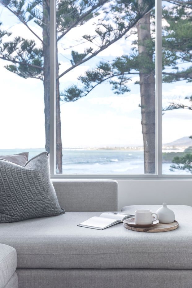 A bright, calm living area featuring a pale grey upholstered sofa or daybed with a textured patterned cushion. A small tray with a cup and ceramic vase rests on the seat beside an open book. Large white-framed windows provide expansive views of the ocean, shoreline, and coastal trees.
