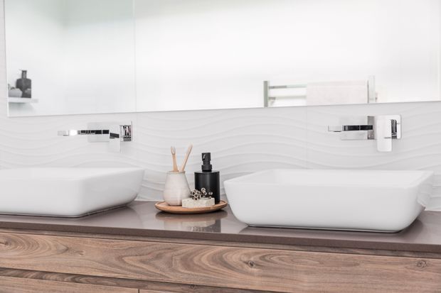 Close-up of a contemporary double bathroom vanity with two white vessel basins, chrome wall-mounted taps, a broad mirror, a white undulating textured tile splashback, and a warm wood vanity cabinet with a dark stone-look benchtop. Minimal bathroom accessories sit between the basins.