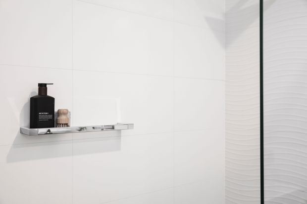 Minimalist white-tiled shower or bathroom wall with a slim chrome floating shelf holding a dark soap dispenser and small toiletry items. A clear glass shower screen with dark edging is visible at right, alongside a white textured wave-pattern wall surface.