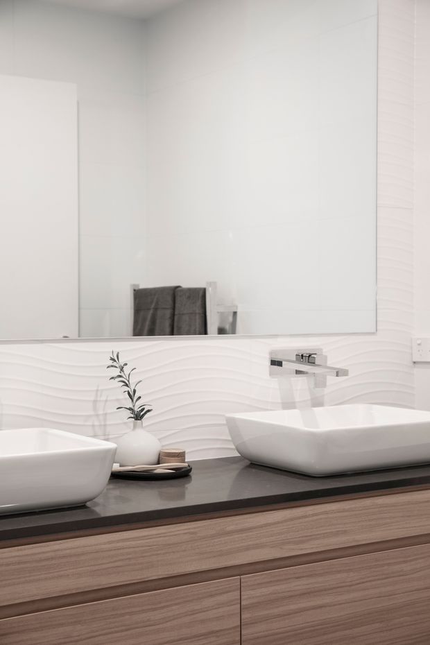 A contemporary bathroom vanity with two white vessel basins on a dark stone countertop, set above warm wood-grain cabinetry. A large mirror, chrome wall-mounted tapware, and white wavy-textured wall tiles create a clean, refined interior.