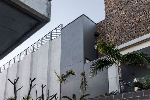 Exterior view of a contemporary multi-level building in a dense urban setting, featuring large smooth grey concrete walls, a dark grey vertical volume, exposed stacked-stone cladding, metal privacy fencing, and tropical palm landscaping. The image is photographed from below, emphasizing the building facade and contrasting material textures.
