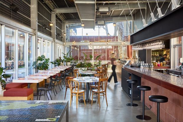 A bright, industrial-style restaurant or café interior with a long bar counter on the right, backbar shelving and hanging glassware, communal and small dining tables, mixed timber and black chairs, and banquette seating. Large glazed openings bring in daylight, while the exposed ceiling services, metal grilles, suspended track lighting, and polished concrete floor reinforce the contemporary hospitality setting.