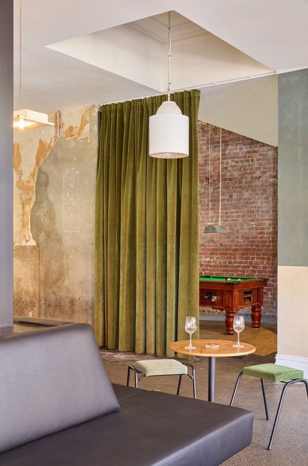 An eclectic interior lounge with a large dark upholstered sofa in the foreground, a small round timber table, green upholstered stools, and a tall olive-green curtain dividing the space. Exposed red brickwork, weathered plaster, concrete-like surfaces, industrial pendant lighting, and a visible vintage-style pool table create a layered industrial character. A large recessed ceiling opening adds architectural interest.