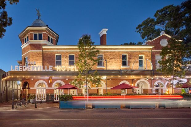 Exterior street view of the Leederville Hotel, a two-storey red-brick heritage-style commercial building with ornate arched openings, decorative white trim, a prominent octagonal corner tower topped with a weathervane, a gabled end with a circular window, and a rooftop sign. Outdoor umbrellas and street-level restaurant seating are visible along the frontage.