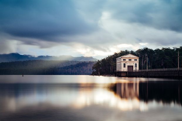 A waterfront architectural exterior showing a small, formal pale masonry building with a symmetrical classical facade and shallow pitched roof, positioned beside a calm lake. Dark forested hills and dramatic overcast mountains form the background, with strong reflections across the water.