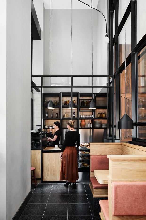 A tall, modern restaurant or café interior viewed from the dining area toward an open service counter and bar. The space features a dramatic double-height volume, black steel-framed glazing and partitions, suspended pendant lights, dark tiled flooring, timber cabinetry and shelving, stainless-steel kitchen equipment, and built-in timber banquette seating upholstered in muted terracotta pink. Two staff members are visible behind the counter. The composition has a refined industrial character with strong vertical lines and warm timber accents.