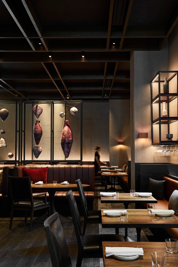 A dimly lit, upscale restaurant interior with dark timber and exposed structural ceiling framing, concrete-toned walls, leather banquette seating, timber dining tables, black chairs, and warm concealed and pendant-style lighting. A sculptural glass display with suspended vessels forms a feature backdrop, while open shelving and a bar area with hanging glassware occupy the right side.