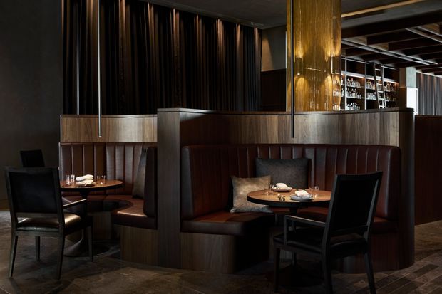 A dark, sophisticated restaurant dining area with curved built-in leather banquettes, round wooden tables, black dining chairs, timber-toned partitions, a visible bar with glassware, and dramatic warm pendant lighting. The space features vertical dark wall detailing and a coffered ceiling, creating an intimate hospitality interior.