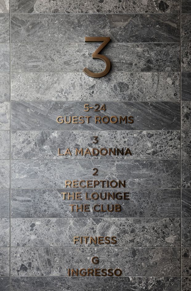 Close-up photograph of a grey, heavily veined and textured stone wall used as a wayfinding directory. Raised bronze or brass-gold lettering and numbers identify guest rooms, La Madonina, reception, lounge, club, fitness, and entrance. The composition is architectural signage-focused with a refined, hotel-like character.