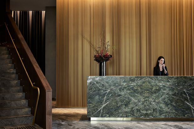 A dramatic, high-end reception lobby with a long stone reception desk in the foreground, backed by a richly textured vertical timber feature wall. A dark stairway with timber handrails and subtle integrated lighting rises at left, while a floral arrangement and receptionist add scale. The space uses dark, moody tones and luxurious natural materials.