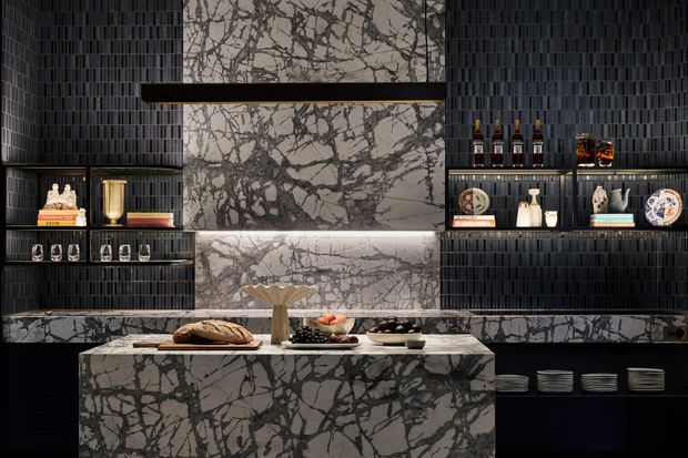 A dramatic, dark-toned contemporary kitchen and entertaining space centred on a large marble island. Black vertically ribbed wall surfaces and integrated open shelving frame a highly veined white-and-grey marble backsplash and island, with concealed linear lighting, glassware, bottles and display objects arranged in the shelving. The composition has a luxurious, moody and highly refined aesthetic.