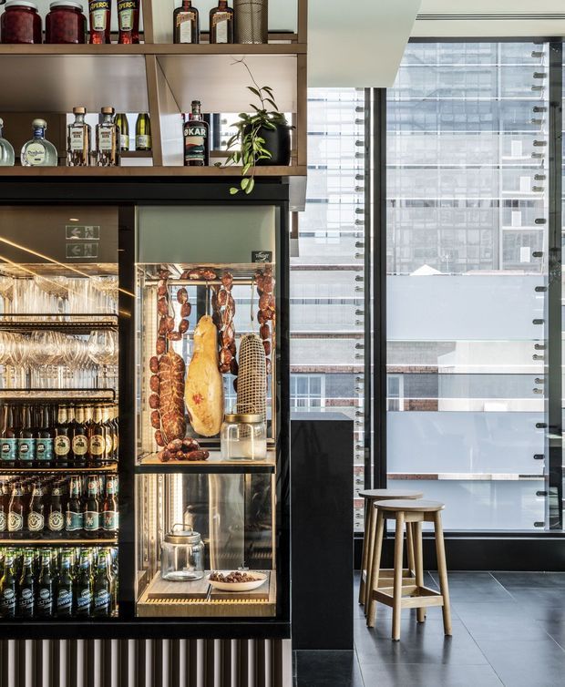 A refined commercial bar or hospitality interior with a dark built-in drinks refrigerator and display cabinet, open timber shelving stocked with bottles and jars, hanging glassware, and a central food display. Floor-to-ceiling glazing provides an urban city view, while two timber stools sit beside a dark tiled floor. The fit-out combines black cabinetry, timber shelving, brushed metal details, and warm integrated lighting.