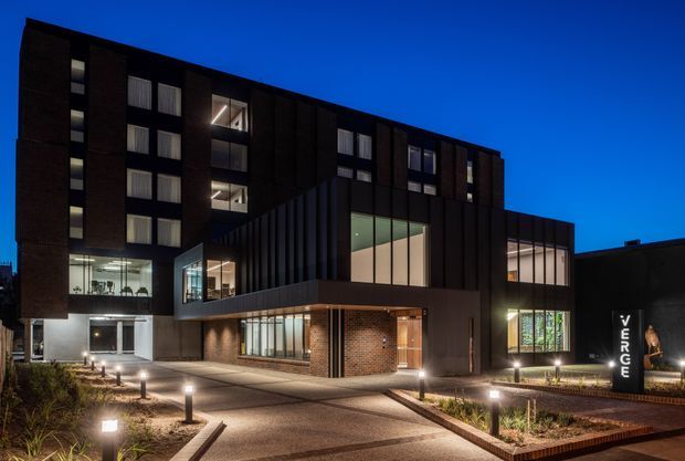 Night-time exterior photograph of a contemporary multi-storey commercial or mixed-use building. The dark brick and vertically ribbed dark facade frame extensive illuminated glazing, recessed balconies, and a projecting lower volume. A landscaped paved forecourt with low bollard lighting leads to the entrance, with a visible 'VERGE' sign on the right.
