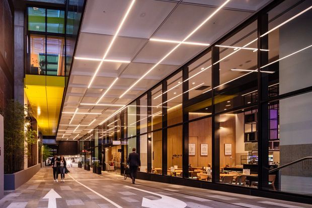 A covered urban pedestrian arcade beside a modern commercial building, with floor-to-ceiling glazed shopfronts, warm timber interiors, dark metal framing, and a dramatic linear illuminated ceiling. People walk along the paved passage, while restaurants or hospitality spaces are visible behind the glazing.