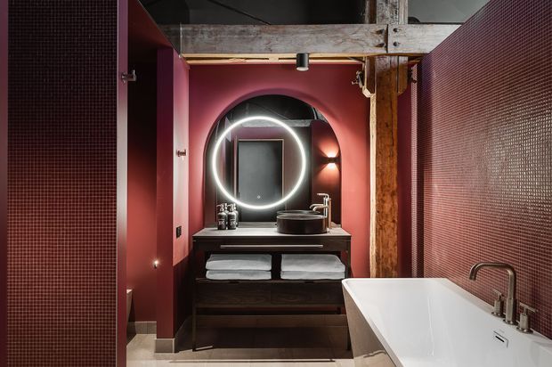 Moody, compact bathroom with a deep burgundy palette, dark wood vanity, round illuminated mirror, black vessel basin, freestanding white bathtub, and exposed rustic timber and beam details. Small-format dark mosaic tile covers the side walls, while warm integrated lighting creates a dramatic, intimate atmosphere.