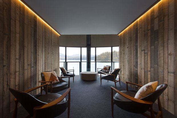 A dark, refined lounge or living room with a symmetrical corridor-like layout. Full-height rustic timber wall panelling lines both sides, each topped with concealed warm linear lighting. Several mid-century-style leather and timber lounge chairs are arranged around a low round ottoman on a dark carpet. Floor-to-ceiling glazing at the far end opens to a calm waterfront or landscape view, with a central dark structural column and a simple charcoal ceiling.