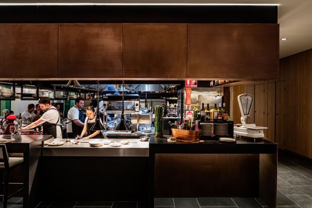 A sophisticated restaurant interior viewed from the dining side toward an open commercial kitchen and service counter. Dark timber or bronze-toned overhead cabinetry, a black counter, stainless-steel cooking equipment, shelving with glassware and bottles, and dark large-format floor tiles create a refined, moody hospitality setting. Chefs and kitchen staff are visible working behind the counter.