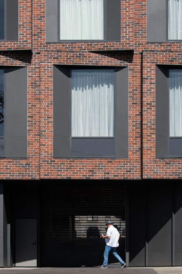Close-up exterior view of a contemporary multi-storey brick building. The facade features repeated dark-framed windows with sheer curtains, projecting brick reveals and deep horizontal overhangs, above a dark recessed ground-level entry or garage opening. A pedestrian provides scale.