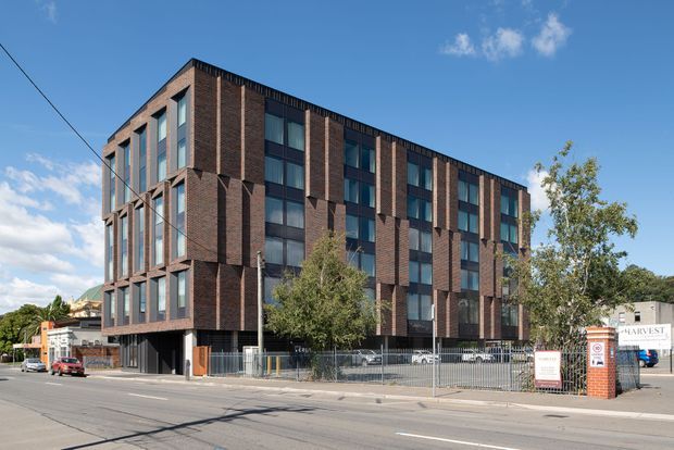 Street-level exterior view of a contemporary multi-storey brick-clad building with a strong vertical facade rhythm, tall dark-framed windows, projecting brick fins, and a fenced entrance forecourt. The urban streetscape includes neighbouring commercial buildings and clear blue sky.