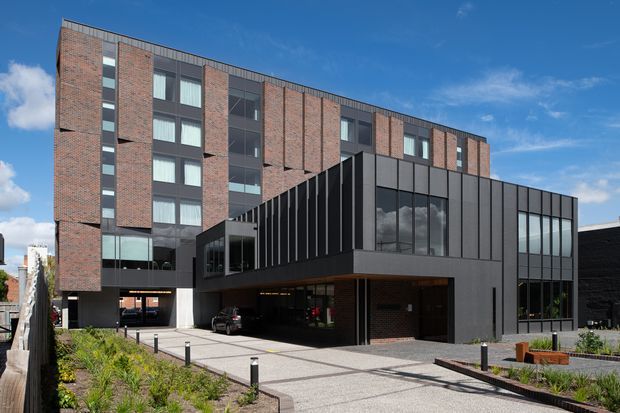 Exterior view of a contemporary multi-storey urban building combining red brick upper levels with dark metal-clad lower volumes and extensive glazing. A paved forecourt with bollards and landscaped planting leads to a covered vehicle entrance and ground-floor windows. Clear blue sky and neighbouring urban buildings are visible.