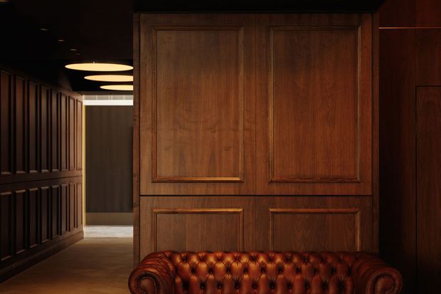 A dark, richly detailed interior hallway or lounge area featuring extensive dark timber wall panelling and a large paneled timber partition or door. Warm circular ceiling lights illuminate a deep brown leather Chesterfield sofa in the foreground, creating a moody, luxurious and traditional atmosphere.