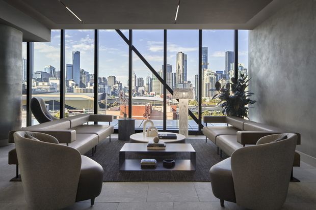 A refined high-rise living room or lounge with expansive floor-to-ceiling windows overlooking a dense city skyline. The space features symmetrical neutral-toned sofas and sculptural armchairs arranged around a low dark coffee table, a large area rug, dark timber or stone flooring, concrete-style columns, and a minimalist recessed ceiling with linear strip lighting. A large indoor plant adds an organic accent beside the glazing.