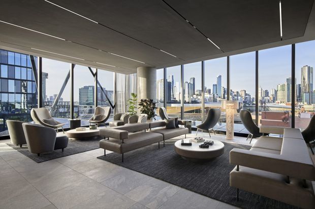 A sophisticated high-rise living room or lounge with floor-to-ceiling glazing overlooking a dense city skyline. The space features a large sectional sofa, sculptural lounge chairs, low round coffee tables, a dark area rug, pale stone-look floor tiles, a concrete structural column, and a dark exposed ceiling with linear integrated lighting. The design is contemporary and refined, with abundant daylight and strong urban views.
