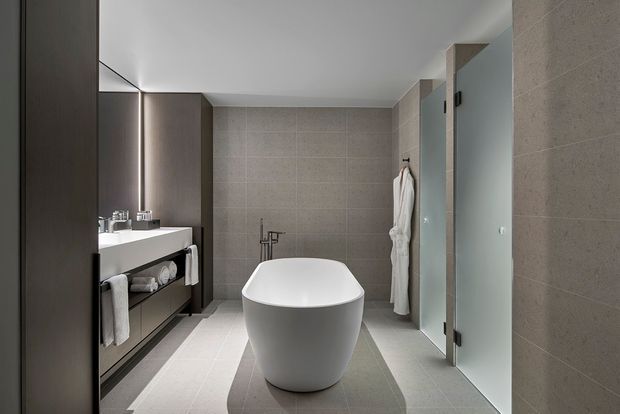 Contemporary bathroom with a freestanding white bathtub centered in the room, a minimalist dark timber vanity with white basin on the left, and a frameless glass shower enclosure on the right. Large-format grey-beige tiles cover the floor and walls, with recessed linear lighting and restrained modern detailing.