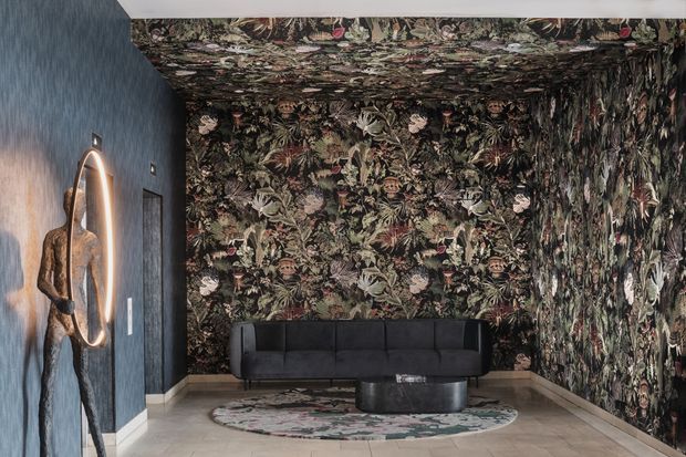 A dramatic, moody lounge or entry seating area with a black upholstered sofa against walls and ceiling covered in dense botanical wallpaper. A sculptural illuminated wall artwork stands on the dark blue textured wall, while a round floral rug and low dark coffee table anchor the space. The room has light stone or tile flooring and a highly decorative, immersive interior finish.