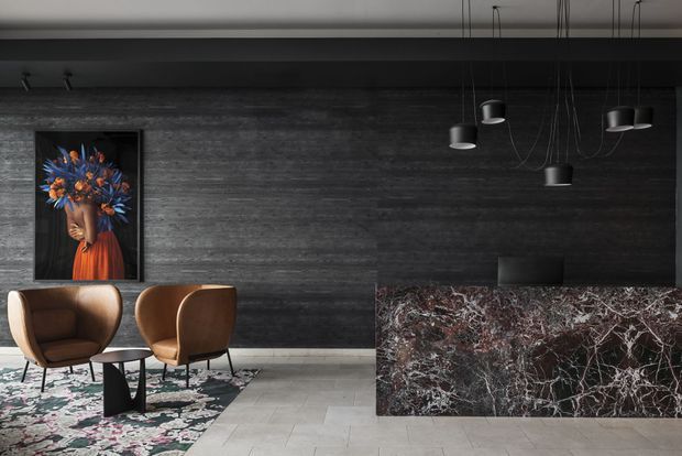 A dramatic contemporary interior featuring a dark textured wall, a striking veined stone reception counter, sculptural black pendant lights, two brown leather lounge chairs, a small black side table, and a bold floral patterned rug. A framed artwork adds a vivid orange and blue accent.