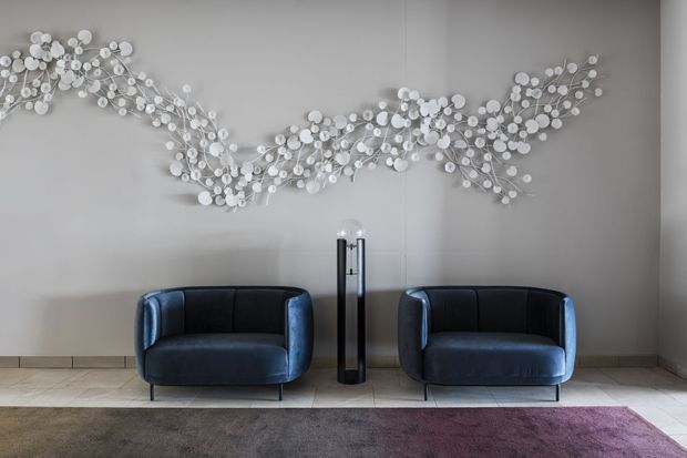 A refined sitting area with two dark blue upholstered armchairs positioned against a warm light-grey wall. A large sculptural white botanical wall installation spans the wall above them, with a slim black floor lamp between the chairs. Pale stone or tile flooring and a muted purple-grey rug complete the calm, contemporary composition.