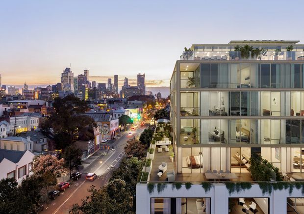 Elevated dusk view of a contemporary multi-storey apartment building with extensive floor-to-ceiling glazing, balconies, rooftop planting and outdoor seating. A dense urban streetscape and illuminated city skyline are visible beside and behind the building.