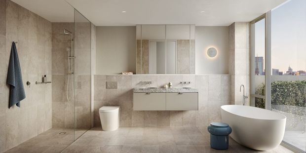 A refined contemporary bathroom with large-format beige stone-look wall and floor surfaces, a frameless glass walk-in shower, wall-hung double vanity with integrated basin, wall-mounted toilet, freestanding oval bathtub, and a narrow blue stool. A full-height window overlooks greenery and a distant urban skyline. The room has recessed ceiling lighting, a round illuminated wall mirror, and a calm neutral palette with a blue accent.