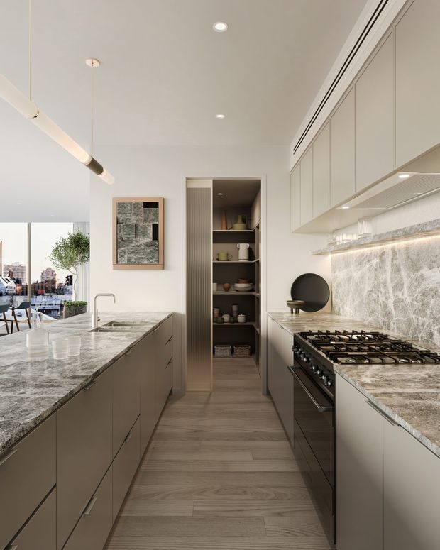 A refined contemporary galley kitchen with a large stone island and matching stone benchtops, integrated pale cabinetry, a full-height veined stone splashback, and a professional gas range. A narrow open pantry with dark shelving sits at the rear, while a glazed dining or living area is visible to the left. Recessed ceiling lights and a linear pendant provide illumination, and timber-look flooring adds warmth.