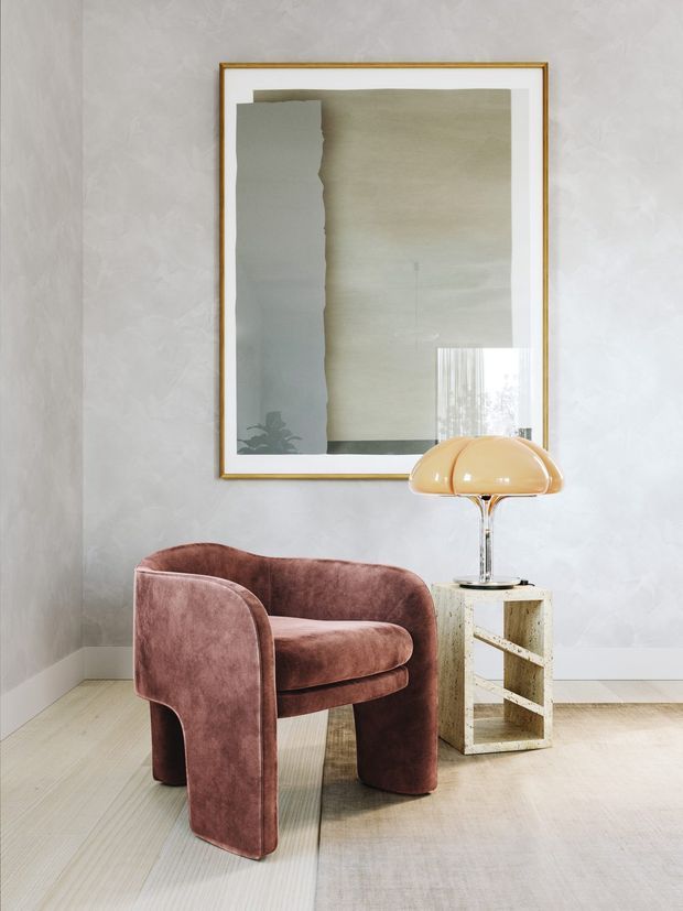 A softly styled interior corner featuring a sculptural mauve velvet armchair beside a small open geometric side table with a warm amber glass lamp. The pale textured plaster walls, light timber flooring, framed abstract artwork, and restrained furnishings create a refined, minimalist setting.