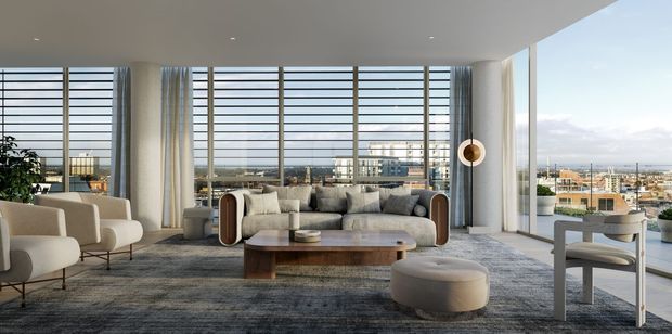 A bright, spacious contemporary living room with expansive floor-to-ceiling glazing, horizontal external sun-control screens, and broad views over an urban or waterfront setting. The room features a large neutral sectional sofa, sculptural lounge chairs, a rounded timber coffee table, a low upholstered ottoman, a textured grey area rug, and a slim modern pendant light. White walls and ceiling, pale timber accents, and abundant natural light create an airy, refined atmosphere.