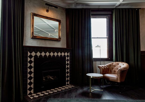 Moody, refined sitting area with a dark timber-panelled fireplace surround, patterned black-and-cream tiled hearth, ornate framed mirror, tall curtained window, tufted leather armchair and small round side table. The room combines traditional detailing with dramatic dark tones and warm leather accents.