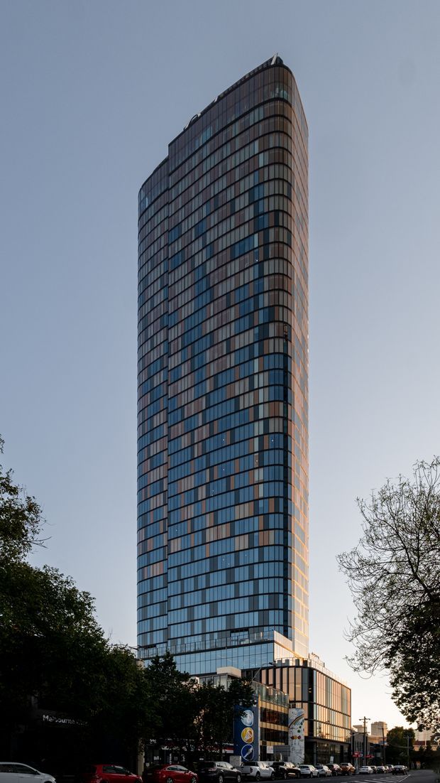 Tall, slender contemporary high-rise tower viewed from street level, with a rounded tapered form, extensive blue-grey curtain-wall glazing, dark horizontal and vertical mullions, and a lower podium containing retail or commercial frontage. Mature trees and parked vehicles frame the urban streetscape.
